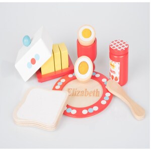 May include: A wooden toy breakfast set with a red and white plate, a wooden knife, a butter dish, a butter knife, a jam jar, and two eggs. The plate has the name "Elizabeth" written on it.