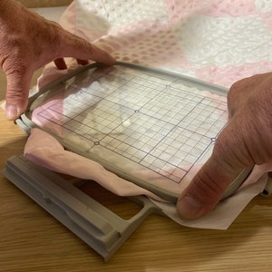 May include: A close-up of a white and pink fabric being placed into a gray embroidery hoop. The hoop has a gridded clear plastic insert.