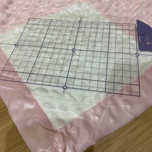 May include: A pink and white quilted blanket with a satin trim. A clear grid ruler is placed on top of the blanket.