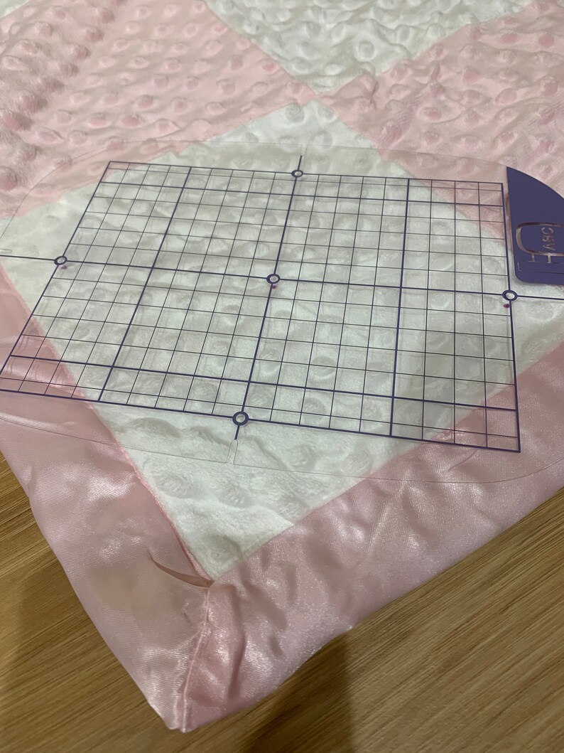 May include: A pink and white quilted blanket with a satin trim. A clear grid ruler is placed on top of the blanket.