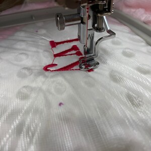 May include: Close-up of a white fabric being embroidered with a red "W" by a sewing machine.