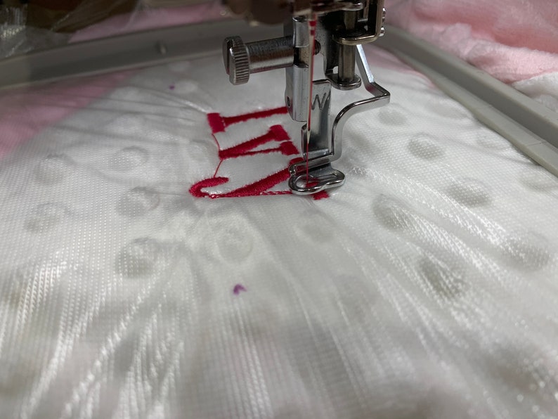 May include: Close-up of a white fabric being embroidered with a red "W" by a sewing machine.