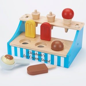May include: A wooden ice cream stand toy with a blue and white striped base. The stand has a sign that reads "Elizabeth's Ice Cream Stand". The toy includes a scoop, a cone, and various ice cream shapes.