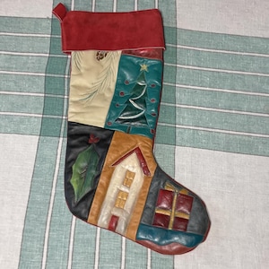 Può includere: Una calza di Natale patchwork con polsino rosso. La calza presenta vari pannelli in pelle con disegni festivi, tra cui un albero di Natale, una casa e regali confezionati. La tavolozza dei colori comprende verde, marrone e rosso.