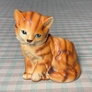 May include: A ceramic figurine of a seated orange tabby cat with black stripes. The cat has blue eyes and a detailed, glossy finish. The figurine is a decorative item, likely for display.