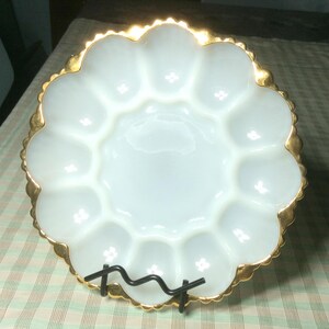 May include: A white, scalloped egg serving dish with a gold-colored trim. The dish has 12 individual egg compartments and is supported by a black metal stand. The dish is made of a glossy material.