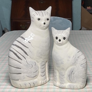 May include: Two ceramic cat figurines. The cats are white with gray stripes and facial details. The larger cat is approximately 12 inches tall, and the smaller cat is about 8 inches tall. They are sitting upright.