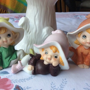Puede incluir: Tres figuritas de porcelana de elfos, cada uno con un sombrero y un atuendo de color diferente. El elfo de la izquierda lleva un sombrero amarillo y un atuendo verde. El elfo del medio lleva un sombrero rosa y un atuendo marrón. El elfo de la derecha lleva un sombrero naranja y un atuendo naranja.