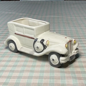 Puede incluir: Una maceta de cerámica blanca rota con forma de coche antiguo. La maceta presenta un diseño de coche clásico con ruedas negras, una franja marrón y faros amarillos. El año "1930" es visible en la parte delantera.