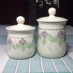 May include: Two off-white ceramic canisters with lids. Each canister features a floral design of purple flowers and green leaves. The canisters have a lattice pattern near the top and a round knob on the lid.