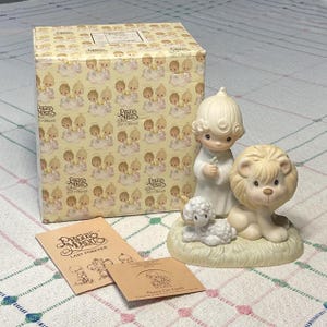 May include: A porcelain figurine featuring a child, a lion, and a lamb on a grassy base, accompanied by a patterned box and two paper inserts. The box and inserts are labeled with the text Precious Moments and the phrase Peace On Earth.