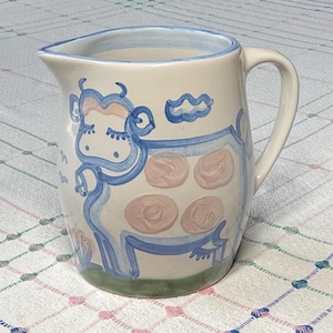 Puede incluir: Jarra de cerámica color crema con un diseño caprichoso de vaca. La vaca está delineada en azul con detalles en rosa y una nube. La jarra tiene un asa curva y un borde azul. La base es verde.