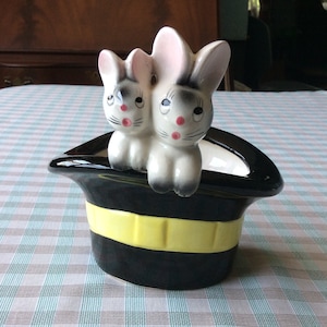 May include: A ceramic planter featuring two white rabbits with gray accents, pink ears, and red noses, emerging from a black top hat with a yellow band. The planter is on a checkered surface.