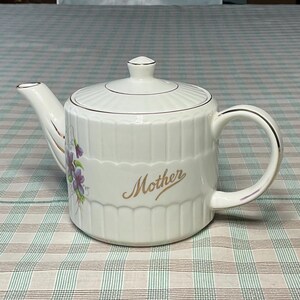 May include: White ceramic teapot with a lid, handle, and spout. The teapot has a gold trim and the word "Mother" in gold script. A floral design with purple flowers and green leaves is on the side. The teapot is on a checkered tablecloth.