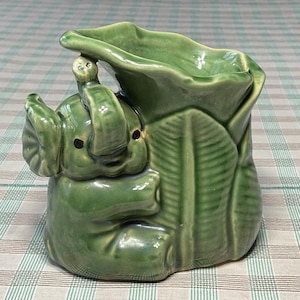 Puede incluir: Una maceta de cerámica verde con forma de elefante. El elefante sostiene una hoja grande, con la trompa enrollada. La maceta tiene un acabado brillante y está sobre una superficie a cuadros.