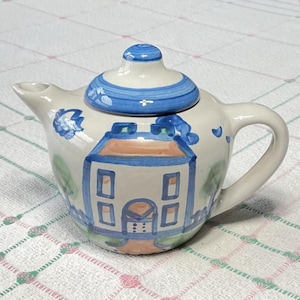 May include: A small, hand-painted ceramic teapot with a whimsical house design. The teapot features a white base with blue and orange accents, including a house illustration on the front and a blue lid. The handle and spout are white.