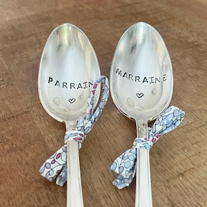 Può includere: Due cucchiai d'argento con fiocchi di nastro blu e rosa. I cucchiai sono incisi con le parole "PARRAIN" e "MARRAINE" con un simbolo di cuore sotto ogni parola.