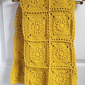 May include: Mustard yellow crochet scarf with a floral pattern, hanging on a wooden hanger. The scarf is made of square crochet blocks, each featuring a central flower design. The scarf is approximately 60 inches long.