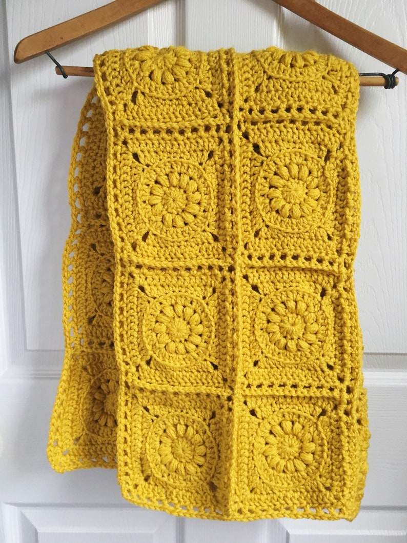 May include: Mustard yellow crochet scarf with a floral pattern, hanging on a wooden hanger. The scarf is made of square crochet blocks, each featuring a central flower design. The scarf is approximately 60 inches long.