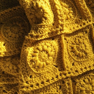 May include: Close-up of a mustard yellow crocheted blanket. The blanket is made of individual square motifs, each featuring a central floral design. The texture is dense and the yarn appears soft. The blanket is folded, creating shadows and depth.
