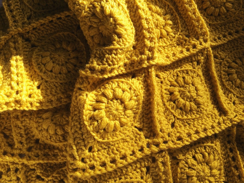 May include: Close-up of a mustard yellow crocheted blanket. The blanket is made of individual square motifs, each featuring a central floral design. The texture is dense and the yarn appears soft. The blanket is folded, creating shadows and depth.