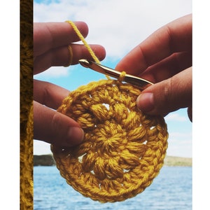 May include: A close-up of a crocheted yellow circle with a crochet hook and yarn. The text "helio square pattern" is at the top. The background shows a blue sky and water.