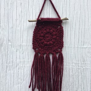 May include: A handmade burgundy crochet wall hanging. The square design features a central floral pattern and long fringe. A wooden dowel and a crocheted loop are used for hanging. The wall hanging is displayed against a white wooden background.