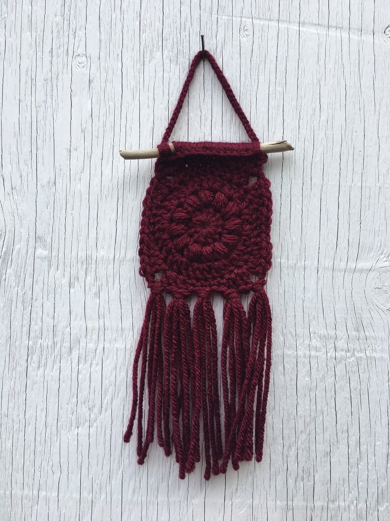 May include: A handmade burgundy crochet wall hanging. The square design features a central floral pattern and long fringe. A wooden dowel and a crocheted loop are used for hanging. The wall hanging is displayed against a white wooden background.