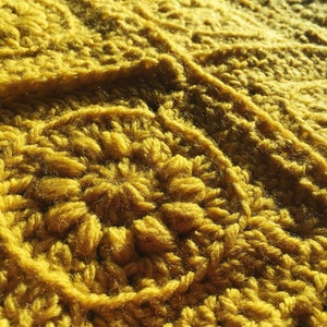 May include: Close-up of a mustard yellow crocheted blanket. The blanket features a textured, raised pattern with a dense, woven appearance. The yarn is thick and the stitches are clearly defined, creating a warm and inviting texture.