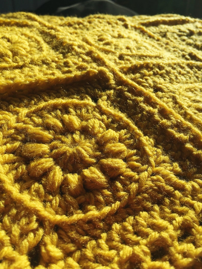May include: Close-up of a mustard yellow crocheted blanket. The blanket features a textured, raised pattern with a dense, woven appearance. The yarn is thick and the stitches are clearly defined, creating a warm and inviting texture.