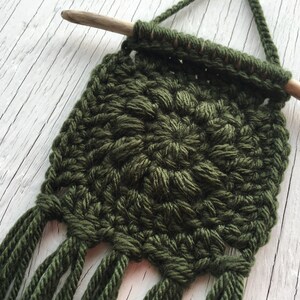 May include: A handmade, olive green macrame wall hanging. The square design features a textured, woven pattern with a wooden dowel at the top for hanging. The bottom edge has several long fringe tassels. This decorative piece adds a touch of bohemian style.