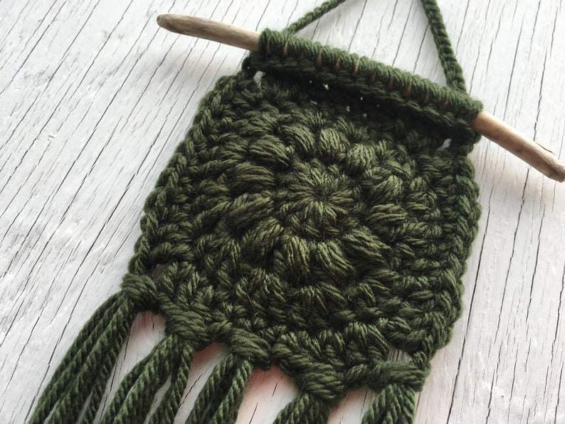 May include: A handmade, olive green macrame wall hanging. The square design features a textured, woven pattern with a wooden dowel at the top for hanging. The bottom edge has several long fringe tassels. This decorative piece adds a touch of bohemian style.