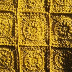 May include: Close-up of a handmade crochet blanket in a mustard yellow color. The blanket is made of individual square motifs, each featuring a central floral design. The squares are joined together with visible seams, creating a textured surface.