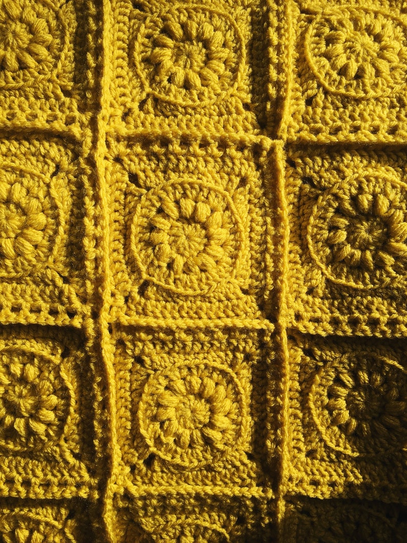 May include: Close-up of a handmade crochet blanket in a mustard yellow color. The blanket is made of individual square motifs, each featuring a central floral design. The squares are joined together with visible seams, creating a textured surface.