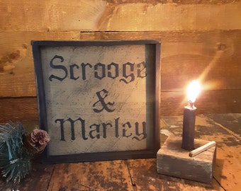 May include: A rustic wooden sign with the words "Scrooge & Marley" in black lettering. A lit candle in a wooden holder sits beside the sign. A small pine branch and pinecone are in the lower left corner. The background is a wooden wall.