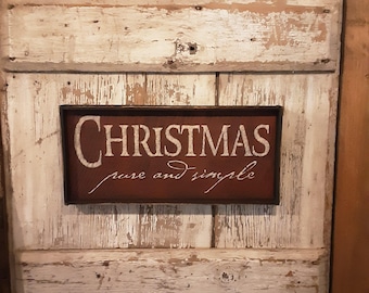 May include: A rustic wooden sign with the word "CHRISTMAS" in white script, framed in a dark brown border. The sign also includes the phrase "pure and simple" in a smaller, elegant font. The background is a distressed, white-painted wooden door.