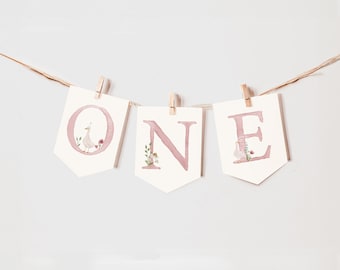 May include: A pink and white banner with the word "ONE" in large letters. Each letter is decorated with a watercolor design of a small bird and flowers.