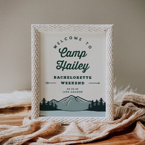 Op de afbeelding: Een wit ingelijst bord met de tekst "Welcome to Camp Hailey Bachelorette Weekend 09.20.30 Lake Amador" in groene tekst. Het bord toont een bergketen met dennenbomen op de achtergrond.