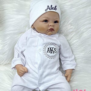 May include: A white baby onesie with a gray wreath embroidered with the letters "AKS" and the name "Alek" on a white beanie.