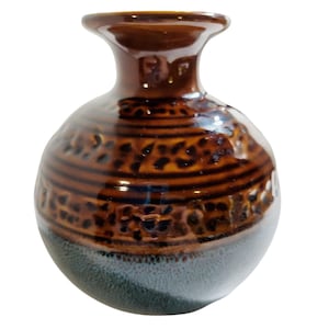 May include: A small, brown ceramic vase with a glossy finish. The vase features a rounded body with a textured, patterned design in a darker brown hue. The base is a muted blue-gray color, and the opening is narrow.