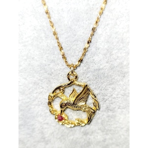 May include: A gold-colored necklace featuring a hummingbird pendant. The pendant is encircled by a floral design and includes a small red gemstone. The chain is a delicate, linked design.
