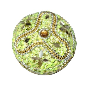 May include: A round, lime green decorative object covered in sequins and embellished with gold and silver beads. The object has a central gold-colored accent and silver mirror-like embellishments. The overall design is ornate and detailed.