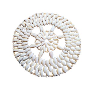 May include: Circular coaster made from white seashells, arranged in a decorative pattern. The shells are tightly packed, creating a textured surface. The coaster is designed to protect surfaces from heat and moisture.
