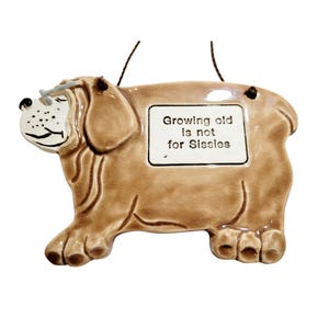 May include: A ceramic dog-shaped wall hanging in shades of brown, with a white rectangular sign that reads "Growing old is not for Sissies." The dog has a smiling face and a wire for hanging.
