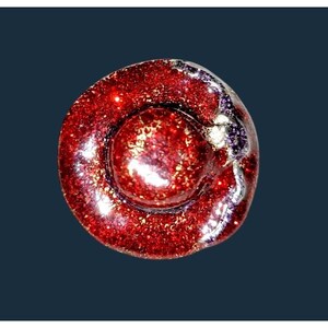 May include: A circular button featuring a vibrant red glitter finish with a raised central dome. The surface displays a reflective, sparkling texture throughout, accented by a small, dark purple and silver metallic detail on the right side.