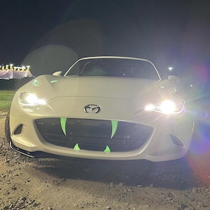 May include: A white Mazda sports car with green fangs on the front grill, illuminated by headlights. The car is parked on a dirt road at night, with a dark sky and distant lights in the background.