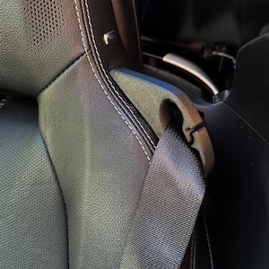 May include: Close-up of a black leather car seat with white stitching. A gray seatbelt is buckled. The seat has a speaker and a plastic component for the seatbelt.