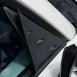 May include: A matte black triangular car window trim piece featuring three small, raised diamond-shaped accents. This textured plastic component is mounted on the exterior corner of a white vehicle near the side mirror and window frame.