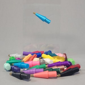 Può includere: Una collezione di punte in plastica colorate per una penna 3D. Le punte sono disposte in un mucchio su una superficie grigia. Una punta blu galleggia sopra il mucchio.