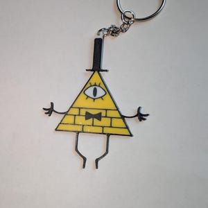 May include: A yellow triangle keychain with a black outline, featuring a single eye, a black bow tie, and a top hat. The keychain has a silver ring and chain. The triangle has a brick-like pattern.